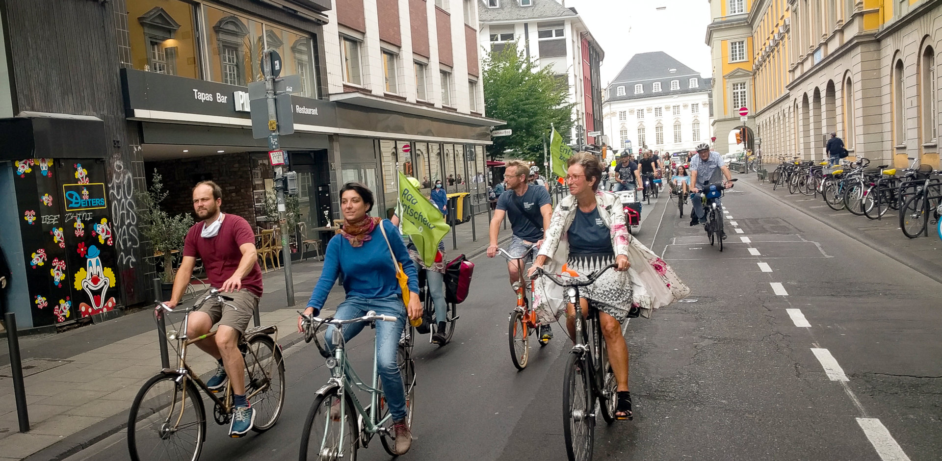 Ein Verband von Radfahrendnen fährt am Uni-Hauptgebäude vorbei. Manche haben Fahnen vom Radentscheid Bonn an ihren Fahrrädern.
