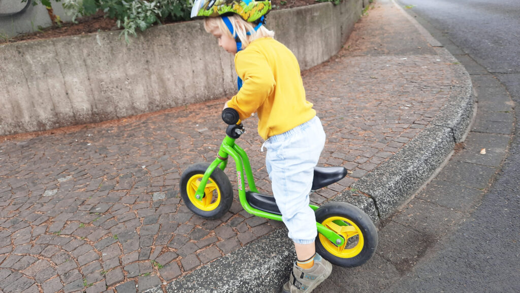 Ein Kind versucht mit seinem Laufrad über einen hohen Boardstein auf den Gehweg zu kommen